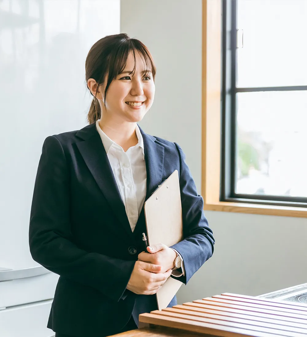 バインダーを抱えて窓際に微笑み立っている女性社員の写真